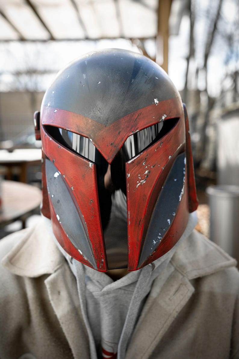 Peut inclure: Casque Mandalorien rouge et noir avec un aspect vieilli. Le casque pr&eacute;sente une visi&egrave;re en forme de T et est port&eacute; sur un sweat &agrave; capuche gris et un manteau clair. Le design du casque comprend des accents gris et noirs.