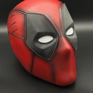 Könnte beinhalten: Eine rot-schwarze Deadpool-Maske mit strukturierter Oberfläche. Die Maske hat schwarze Augenabdeckungen mit weißen Netzkonturen. Die Maske ist eine Nachbildung der Maske der Figur aus den Marvel Comics.
