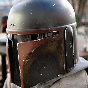 Peut inclure: Un casque Mandalorien noir et rouge avec un aspect vieilli. Le casque a un dessus arrondi et une visi&egrave;re en forme de T. La palette de couleurs comprend des accents noirs, rouges et argent&eacute;s, avec des signes d'usure visibles.
