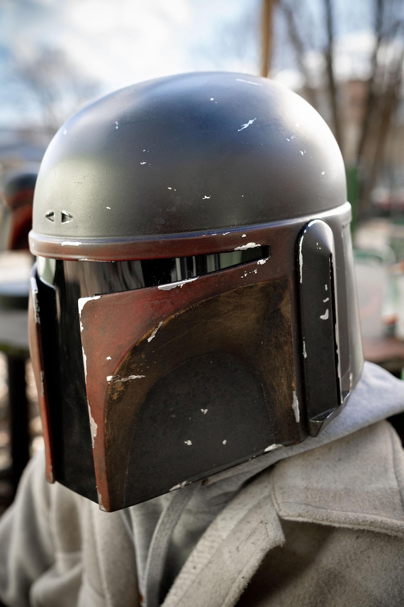Peut inclure: Un casque Mandalorien noir et rouge avec un aspect vieilli. Le casque a un dessus arrondi et une visi&egrave;re en forme de T. La palette de couleurs comprend des accents noirs, rouges et argent&eacute;s, avec des signes d'usure visibles.