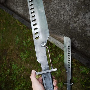 May include: A metallic blade with a series of rectangular cutouts along the spine, featuring a dark rectangular handle and a thin support rod connecting the blade to the hilt. The surface has a weathered, brushed finish with visible scuffs.