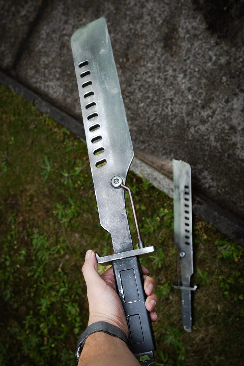 May include: A metallic blade with a series of rectangular cutouts along the spine, featuring a dark rectangular handle and a thin support rod connecting the blade to the hilt. The surface has a weathered, brushed finish with visible scuffs.