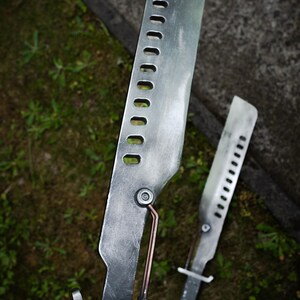 May include: Two silver-colored metal blades with a weathered finish, each featuring a row of rectangular cutouts along the spine and a copper-toned rod connecting the blade to the handle. The handles are black with a prominent crossguard.