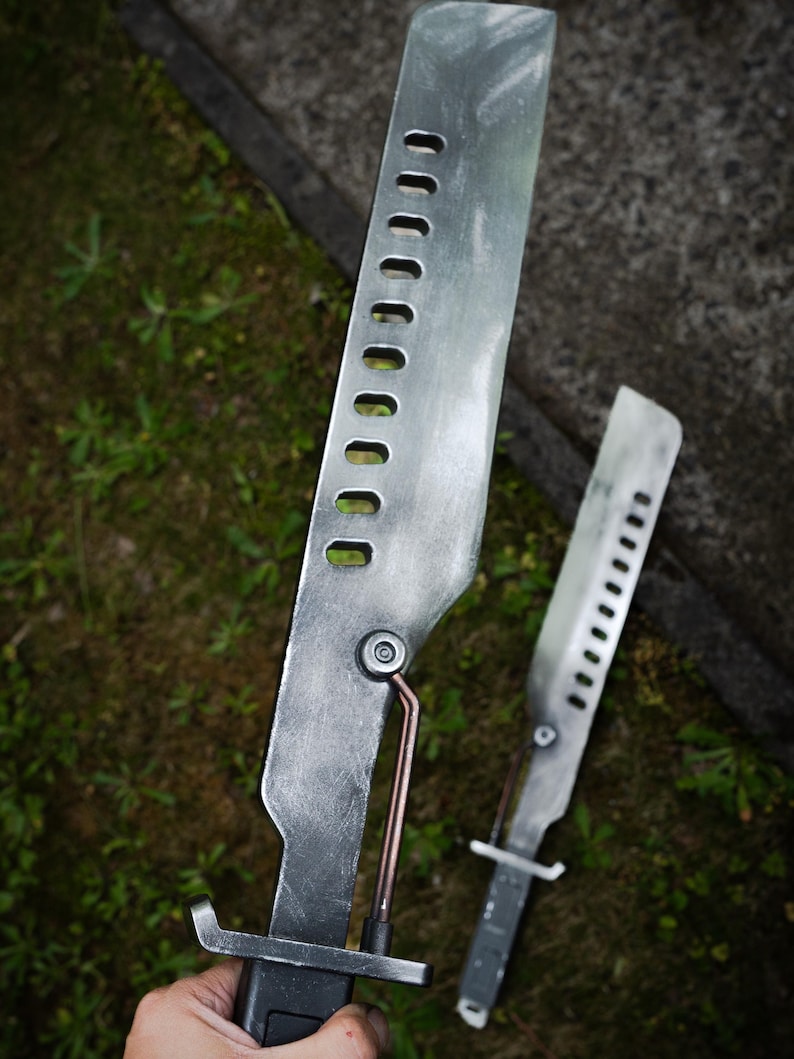 May include: Two silver-colored metal blades with a weathered finish, each featuring a row of rectangular cutouts along the spine and a copper-toned rod connecting the blade to the handle. The handles are black with a prominent crossguard.