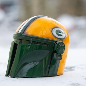 Peut inclure: Casque personnalis&eacute; de style Mandalorien, jaune et vert, avec une bande blanche et un logo "G" vert sur le c&ocirc;t&eacute;. L'objet est pos&eacute; sur un fond enneig&eacute;.
