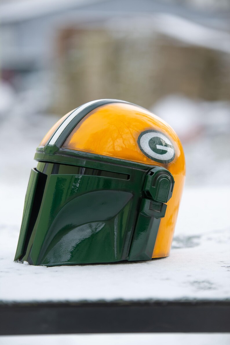Peut inclure: Casque personnalis&eacute; de style Mandalorien, jaune et vert, avec une bande blanche et un logo "G" vert sur le c&ocirc;t&eacute;. L'objet est pos&eacute; sur un fond enneig&eacute;.