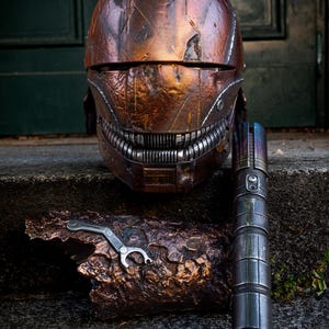 Op de afbeelding: Een verweerde koperkleurige helm met een getextureerd oppervlak en een metalen rooster-mondstuk, vergezeld van een lange, cilindrische donker-metalen handgreep en een klein, zilverkleurig mechanisch onderdeel op een ruwe, bronskleurige basis.