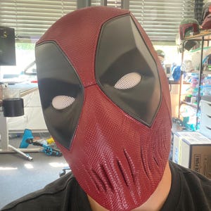 Puede incluir: Una m&aacute;scara de Deadpool roja y negra con una superficie texturizada. La m&aacute;scara tiene un &aacute;rea de ojos negra con ojos de malla blanca.
