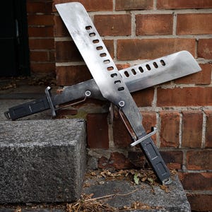 May include: Two metallic machete-style blades with a weathered finish, each featuring a series of rectangular cutouts along the spine. The blades have dark rectangular handles and are crossed, showing a silver-toned metal construction.