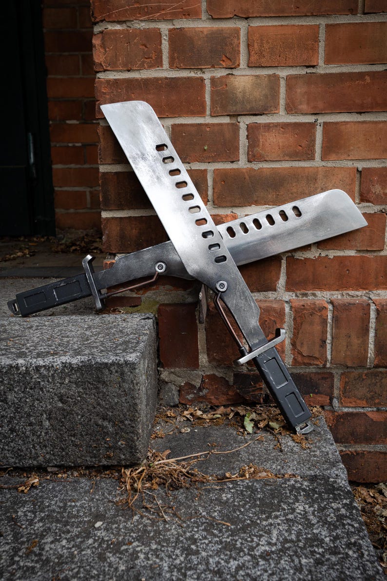 May include: Two metallic machete-style blades with a weathered finish, each featuring a series of rectangular cutouts along the spine. The blades have dark rectangular handles and are crossed, showing a silver-toned metal construction.