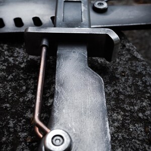 May include: A dark gray metal bayonet with a weathered, scratched finish, featuring a rectangular crossguard, a copper-colored wire element attached to the side, and a cylindrical fastener near the handle, positioned against a matching scabbard.