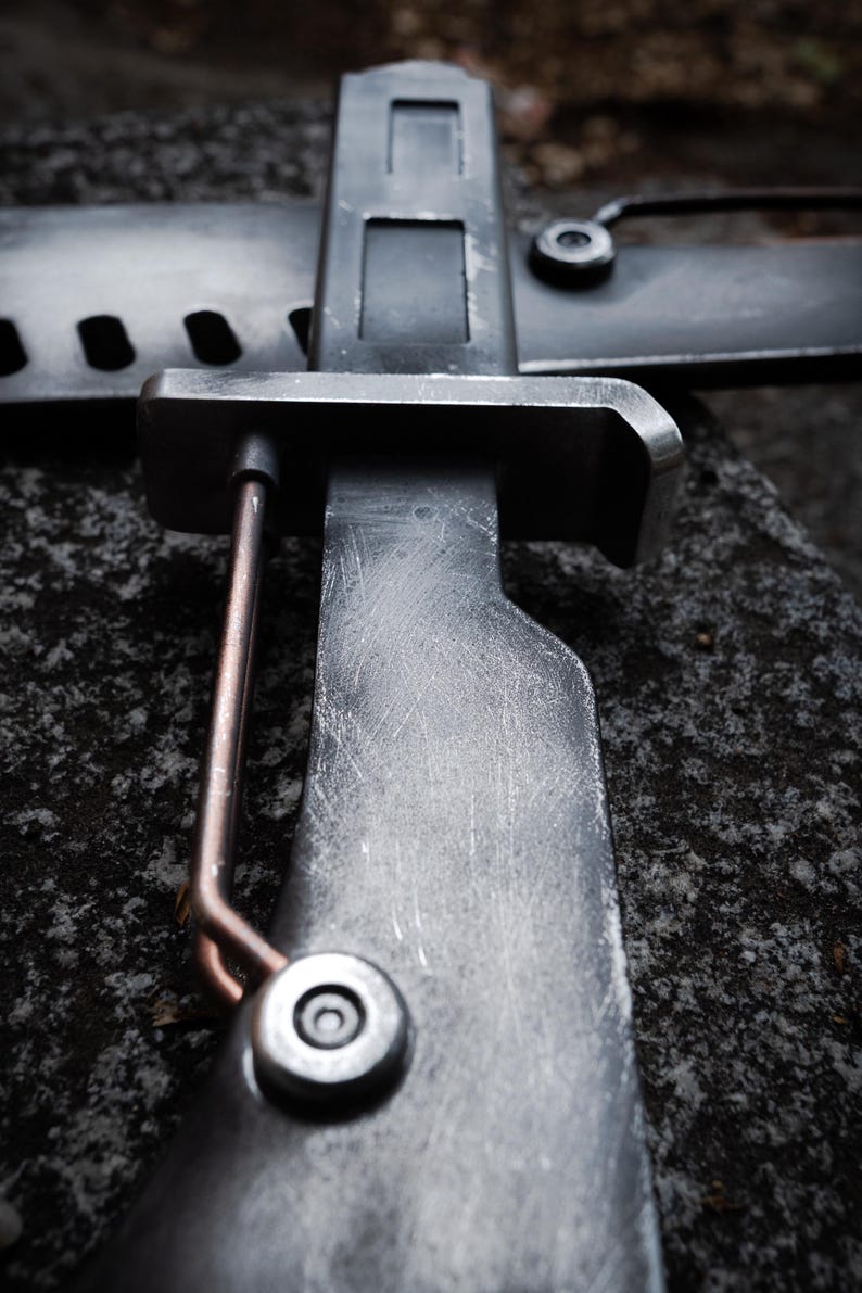 May include: A dark gray metal bayonet with a weathered, scratched finish, featuring a rectangular crossguard, a copper-colored wire element attached to the side, and a cylindrical fastener near the handle, positioned against a matching scabbard.