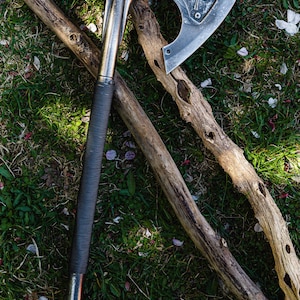 Könnte beinhalten: Eine Metall-Axt mit silberner Klinge und schwarzem Griff, die auf Gras und zwei Holzästen liegt. Der Axtkopf hat ein dekoratives Emblem. Der Griff ist mit einem strukturierten, dunklen Material umwickelt.