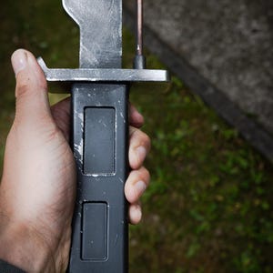 May include: A bayonet with a weathered silver-toned blade and a matte black handle featuring two rectangular recessed panels. The crossguard is silver-toned metal, and a thin copper-colored wire runs vertically along the side of the blade.