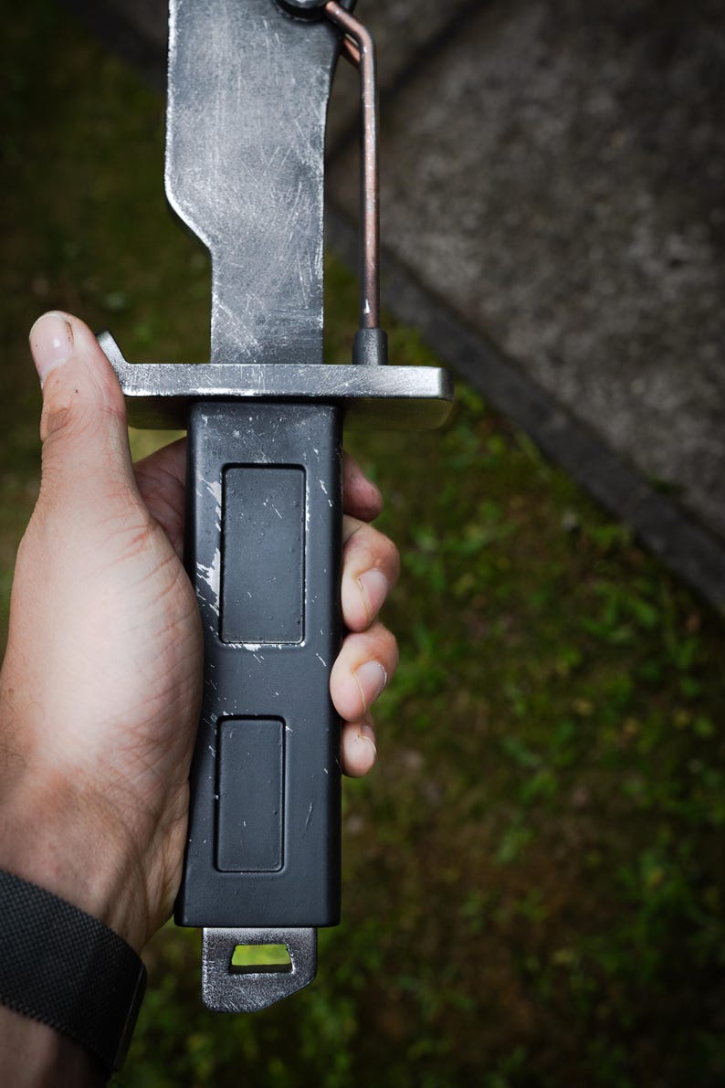 May include: A bayonet with a weathered silver-toned blade and a matte black handle featuring two rectangular recessed panels. The crossguard is silver-toned metal, and a thin copper-colored wire runs vertically along the side of the blade.