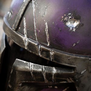 Peut inclure: Gros plan sur un casque violet et argent&eacute; vieilli. Le casque a une finition m&eacute;tallique avec des rayures et une usure visibles, sugg&eacute;rant une esth&eacute;tique us&eacute;e au combat. La conception comprend une visi&egrave;re et un dessus arrondi.