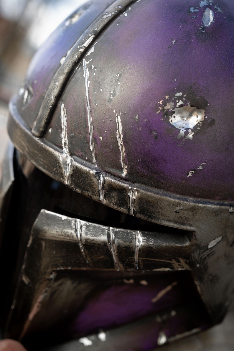Peut inclure: Gros plan sur un casque violet et argent&eacute; vieilli. Le casque a une finition m&eacute;tallique avec des rayures et une usure visibles, sugg&eacute;rant une esth&eacute;tique us&eacute;e au combat. La conception comprend une visi&egrave;re et un dessus arrondi.
