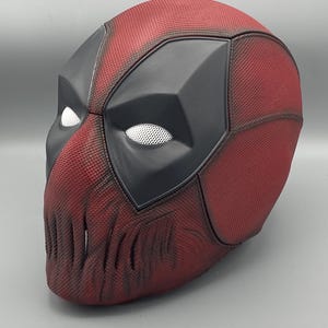 Puede incluir: Una m&aacute;scara de Deadpool roja y negra con una superficie texturizada y un dise&ntilde;o detallado. La m&aacute;scara tiene un &aacute;rea de ojos negra con ojos de malla blanca.