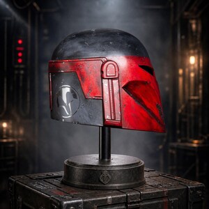 Peut inclure: Un casque Mandalorien, principalement noir et rouge, est pr&eacute;sent&eacute; sur un socle au-dessus d'une bo&icirc;te en m&eacute;tal textur&eacute;. Le casque pr&eacute;sente un aspect vieilli avec une visi&egrave;re et des d&eacute;tails lat&eacute;raux distinctifs. L'arri&egrave;re-plan est un cadre industriel faiblement &eacute;clair&eacute;.