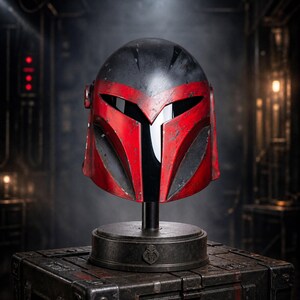Peut inclure: Un casque Mandalorien rouge et noir, expos&eacute; sur un support au-dessus d'une bo&icirc;te en m&eacute;tal. Le casque pr&eacute;sente une visi&egrave;re distinctive en forme de T et se d&eacute;tache sur un fond sombre et industriel. Le design du casque est d&eacute;taill&eacute; et vieilli.