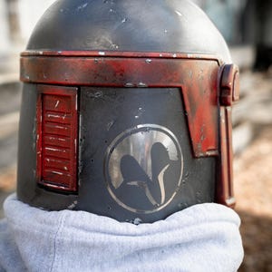 Peut inclure: Casque gris fonc&eacute; vieilli avec des d&eacute;tails rouges, orn&eacute; d'un embl&egrave;me circulaire. Le casque a un dessus arrondi et un panneau lat&eacute;ral rectangulaire. Le casque est port&eacute; sur un sweat &agrave; capuche gris.