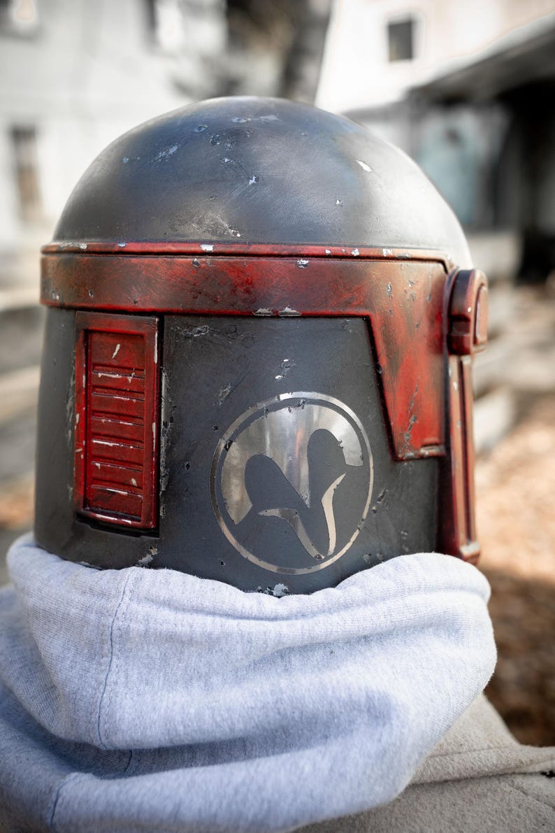 Peut inclure: Casque gris fonc&eacute; vieilli avec des d&eacute;tails rouges, orn&eacute; d'un embl&egrave;me circulaire. Le casque a un dessus arrondi et un panneau lat&eacute;ral rectangulaire. Le casque est port&eacute; sur un sweat &agrave; capuche gris.