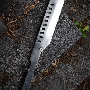 May include: A silver-colored metal machete with a rectangular handle and a long blade featuring a row of ten oval cutouts. A thin metal rod connects the handle to the base of the blade, which has a flat, squared-off tip and a brushed finish.