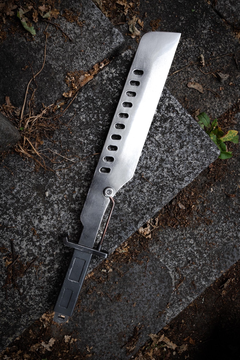 May include: A silver-colored metal machete with a rectangular handle and a long blade featuring a row of ten oval cutouts. A thin metal rod connects the handle to the base of the blade, which has a flat, squared-off tip and a brushed finish.
