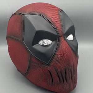 Puede incluir: Una m&aacute;scara de Deadpool roja y negra con una superficie texturizada. La m&aacute;scara tiene una zona de ojos negra con agujeros de ojos de malla blanca y una zona de boca negra con una serie de hendiduras verticales.