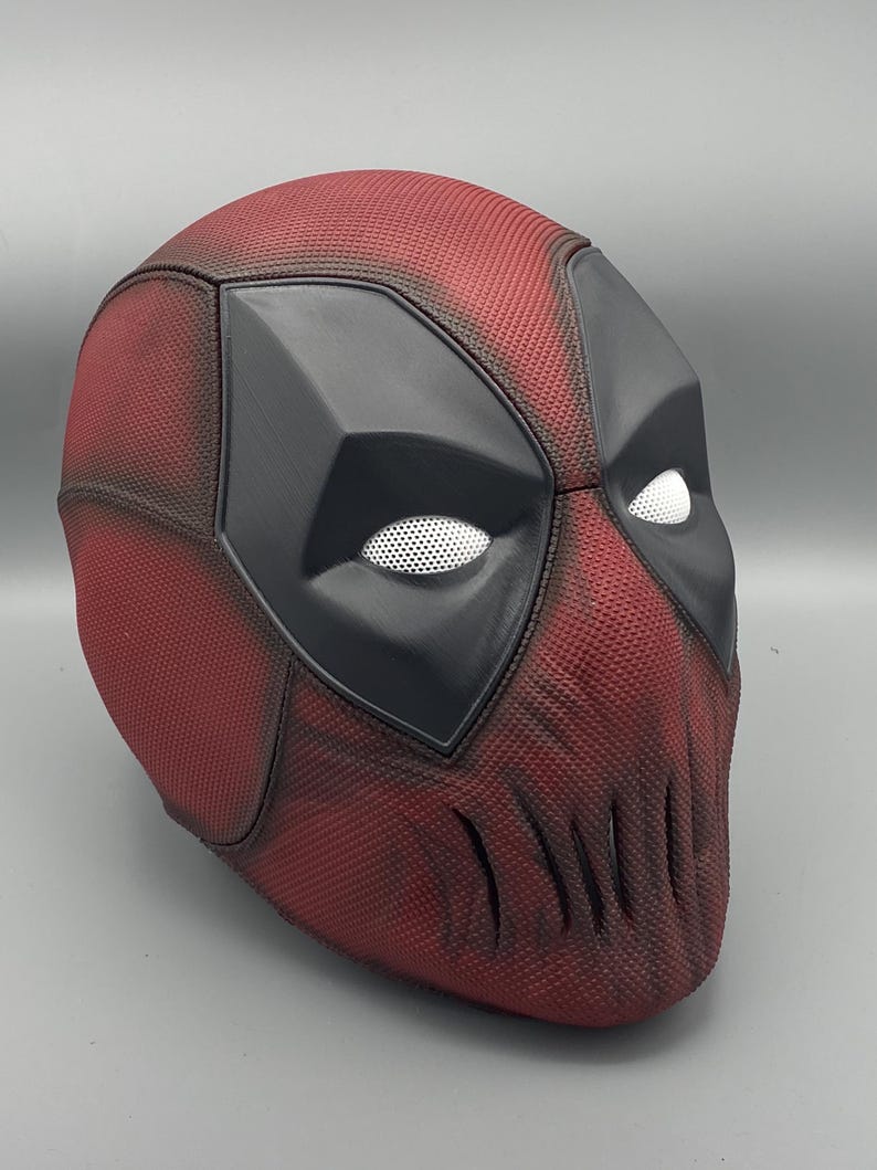 Puede incluir: Una m&aacute;scara de Deadpool roja y negra con una superficie texturizada. La m&aacute;scara tiene una zona de ojos negra con agujeros de ojos de malla blanca y una zona de boca negra con una serie de hendiduras verticales.
