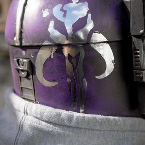 Peut inclure: Gros plan sur un casque violet avec un embl&egrave;me argent&eacute;. Le casque a un aspect vieilli avec des garnitures et des d&eacute;tails argent&eacute;s. L'embl&egrave;me est un motif stylis&eacute; avec des formes courbes. Un sweat &agrave; capuche gris est visible en dessous.