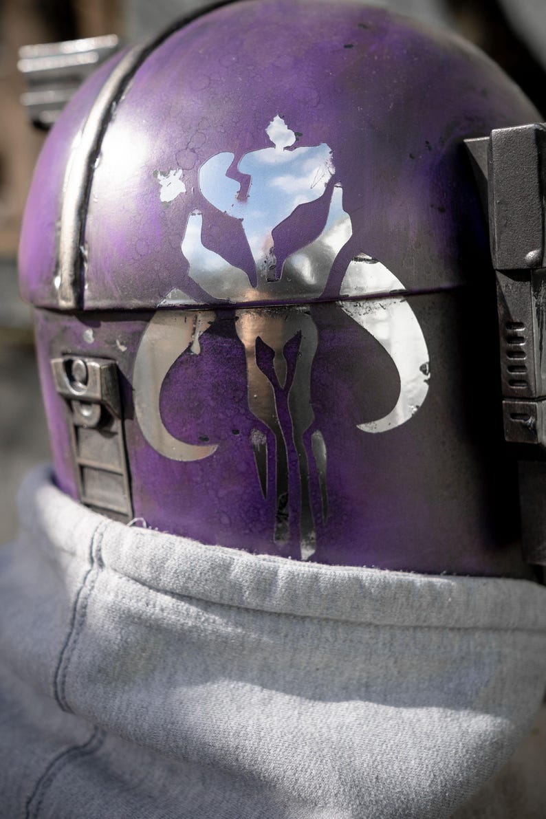 Peut inclure: Gros plan sur un casque violet avec un embl&egrave;me argent&eacute;. Le casque a un aspect vieilli avec des garnitures et des d&eacute;tails argent&eacute;s. L'embl&egrave;me est un motif stylis&eacute; avec des formes courbes. Un sweat &agrave; capuche gris est visible en dessous.