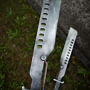 May include: Two metallic, silver-toned blades with a weathered finish, featuring a row of rectangular cutouts along the spine. Each blade has a copper-colored rod connecting the handle to the blade body and a dark, rectangular grip handle.