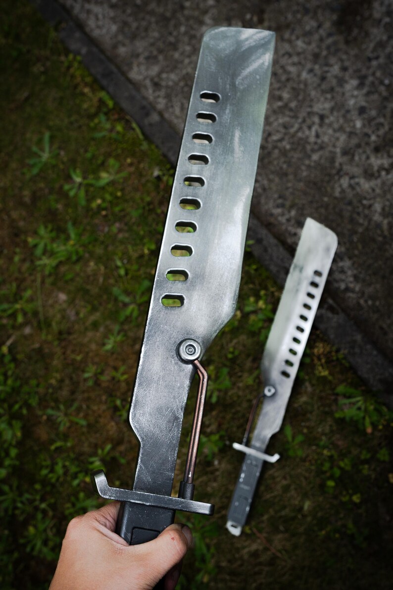 May include: Two metallic, silver-toned blades with a weathered finish, featuring a row of rectangular cutouts along the spine. Each blade has a copper-colored rod connecting the handle to the blade body and a dark, rectangular grip handle.