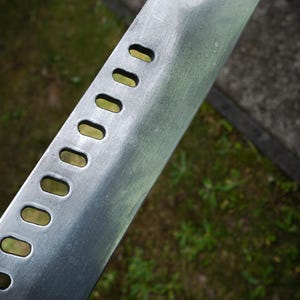 May include: A silver-colored metal blade featuring a series of eight evenly spaced, elongated oval cutouts along the side. The surface of the metal has a brushed, matte finish with visible texture and a slightly angled, tapered edge at the top.
