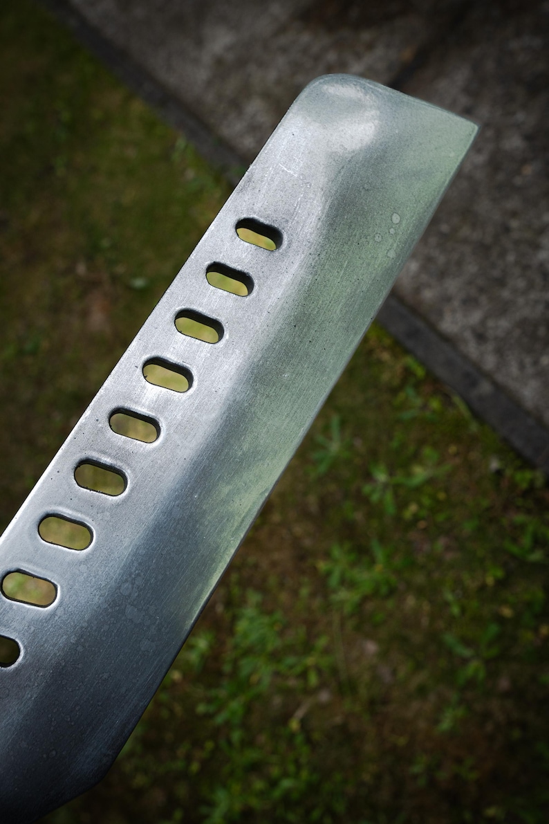 May include: A silver-colored metal blade featuring a series of eight evenly spaced, elongated oval cutouts along the side. The surface of the metal has a brushed, matte finish with visible texture and a slightly angled, tapered edge at the top.