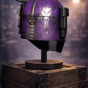 Peut inclure: Un casque violet avec des d&eacute;tails argent&eacute;s et un embl&egrave;me blanc, pr&eacute;sent&eacute; sur un support en bois sur une bo&icirc;te en bois. Le casque a un aspect vieilli, probablement un accessoire ou un objet de collection.