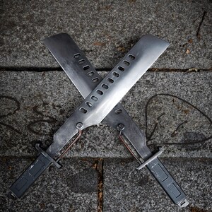 May include: Two metallic blades arranged in an X-shape, each featuring a long, flat edge, a series of rectangular cutouts along the spine, a dark grey handle, and a thin copper-colored wire connecting the handle to the base of the blade.
