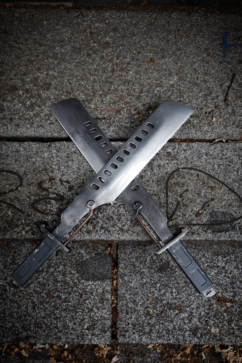 May include: Two metallic blades arranged in an X-shape, each featuring a long, flat edge, a series of rectangular cutouts along the spine, a dark grey handle, and a thin copper-colored wire connecting the handle to the base of the blade.