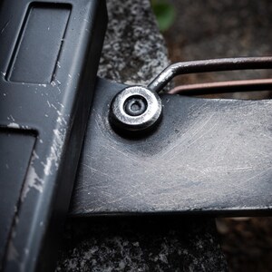 May include: A close-up view of a dark gray metal tool featuring a matte finish with visible surface scratches. The object includes a prominent circular silver-toned rivet or fastener connecting a rectangular handle section to a flat blade-like component.