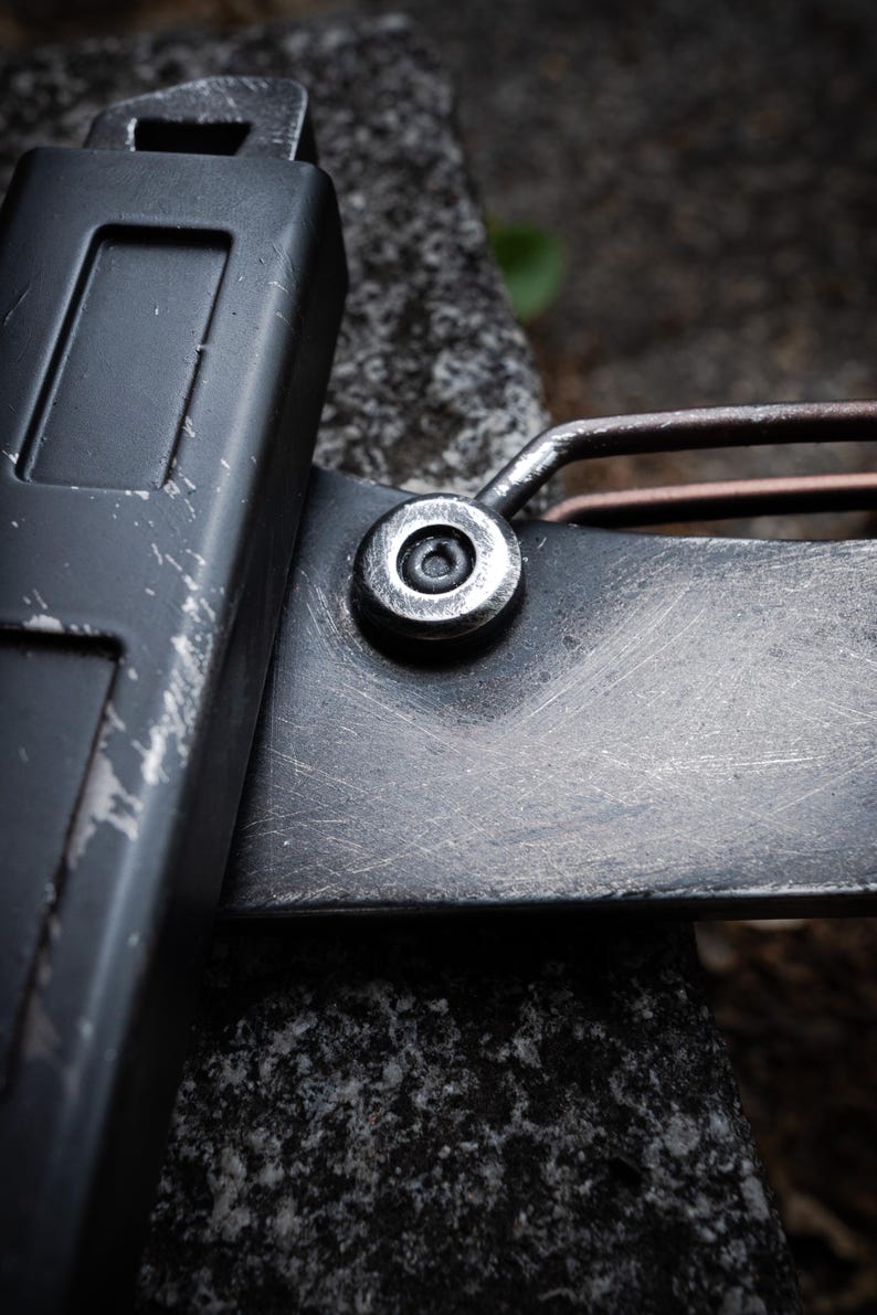 May include: A close-up view of a dark gray metal tool featuring a matte finish with visible surface scratches. The object includes a prominent circular silver-toned rivet or fastener connecting a rectangular handle section to a flat blade-like component.