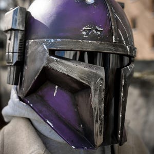 Peut inclure: Gros plan sur un casque Mandalorien violet et noir. Le casque pr&eacute;sente des signes d'usure, avec des rayures visibles. Il est &eacute;quip&eacute; d'une visi&egrave;re et d'un composant lat&eacute;ral.