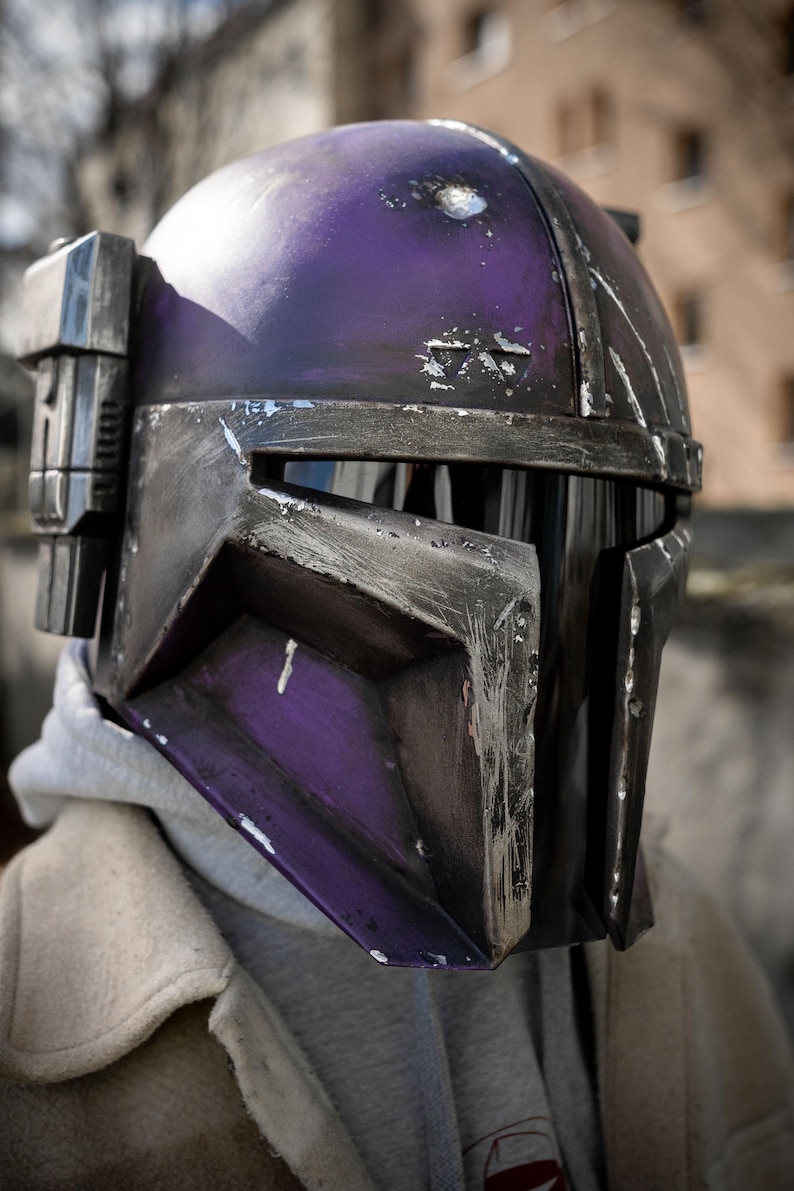 Peut inclure: Gros plan sur un casque Mandalorien violet et noir. Le casque pr&eacute;sente des signes d'usure, avec des rayures visibles. Il est &eacute;quip&eacute; d'une visi&egrave;re et d'un composant lat&eacute;ral.