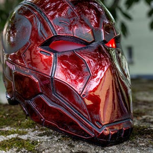 Puede incluir: Un casco rojo y negro brillante con un diseño futurista. El casco presenta paneles angulares, remaches visibles y lentes oculares rojas brillantes. La estética sugiere un tema de ciencia ficción o cosplay.