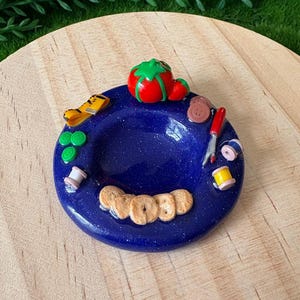 May include: A miniature blue dish with various craft-themed items. The dish contains a red tomato, a yellow tape measure, green circles, brown buttons, a red pen, spools of thread, and small bagels. The dish is on a wooden surface.