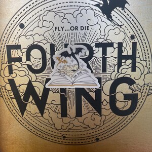 K&ouml;nnte beinhalten: Ein Buchcover mit einem schwarzen und goldenen Design, das die Worte "Fourth Wing" und einen Drachen zeigt. Das Buchcover hat einen Aufkleber mit einem Drachen und den Worten "Fly...or Die" darauf.