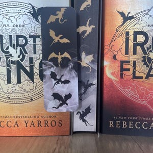 May include: A black bookmark featuring gold and black dragon silhouettes, placed between three books. The books have orange, black, and dark blue covers. The bookmark has a smoky design at the bottom. The books are by Tracy Wolff, Rebecca Yarros, and another author.