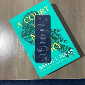 May include: A black bookmark with a celestial design and the text "to the stars who listen and the dreams that are answered". The bookmark is on top of a teal book with the title "A Court of Thorns and Roses" and the author's name "Sarah J. Maas".