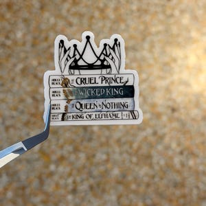 May include: A white sticker featuring a stack of books with the titles "The Cruel Prince", "Wicked King", "Queen of Nothing", and "King of Elfhame". The sticker has a crown design and the author's name, Holly Black.