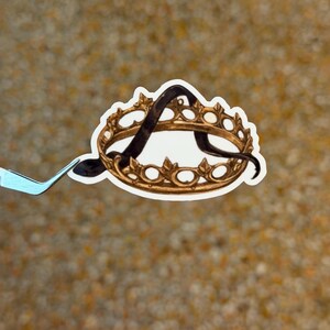 May include: A sticker featuring a gold-colored crown with a black letter "A" inside. The crown has a detailed design with small, circular accents. The sticker is held by a pair of tweezers against a textured, tan background.